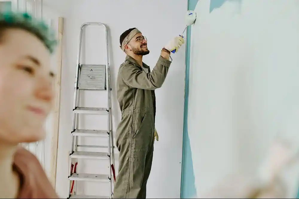 Bir adam, merdivenin yanında duvarı rulo ile boyuyor. Ön planda bulanık bir kişi görülüyor