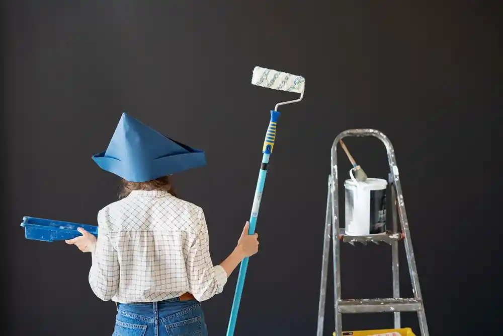 Mavi şapka takan kişi, boya rulosu ve boya tepsisi tutarak merdivenin yanında duruyor