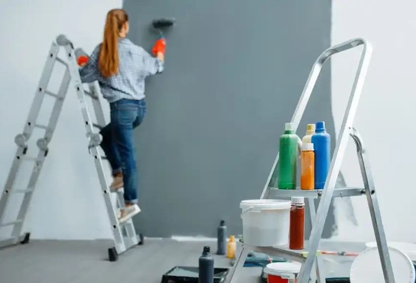 Kadın, merdiven üzerinde duvarı boyarken ön planda boya malzemeleri bulunan bir merdiven duruyor
