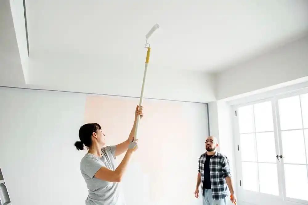 Bir kadın, uzatma saplı boya rulosuyla duvarı boyarken bir adam onu izliyor