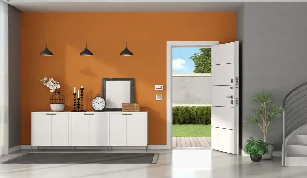 Turuncu duvarlı modern giriş holünde beyaz konsol, üç sarkıt lamba ve açık bir dış kapı görülüyor