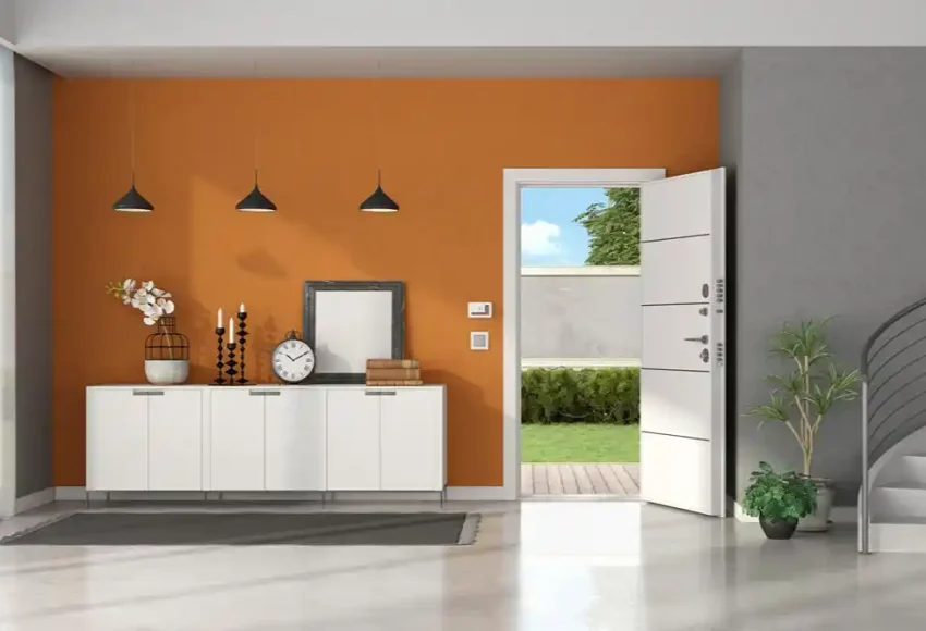 Turuncu duvarlı modern giriş holünde beyaz konsol, üç sarkıt lamba ve açık bir dış kapı görülüyor